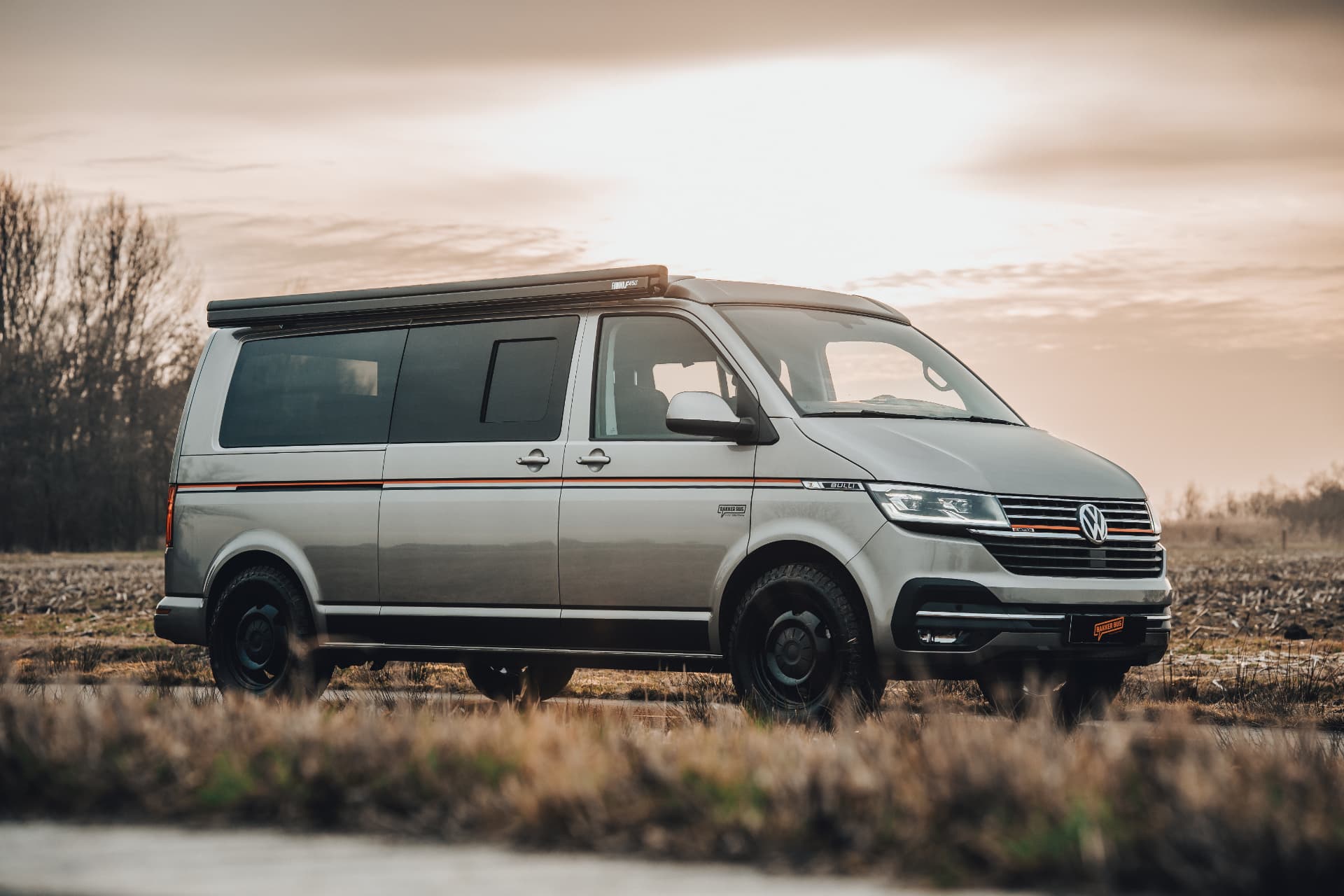 Zilveren VW camperbus in een veld bij zonsondergang